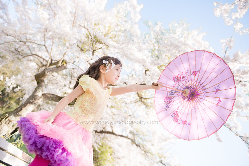 Young girl with pretty purple parasol stands beneath a blossom covered tree. Child photography in Vancouver B.C.