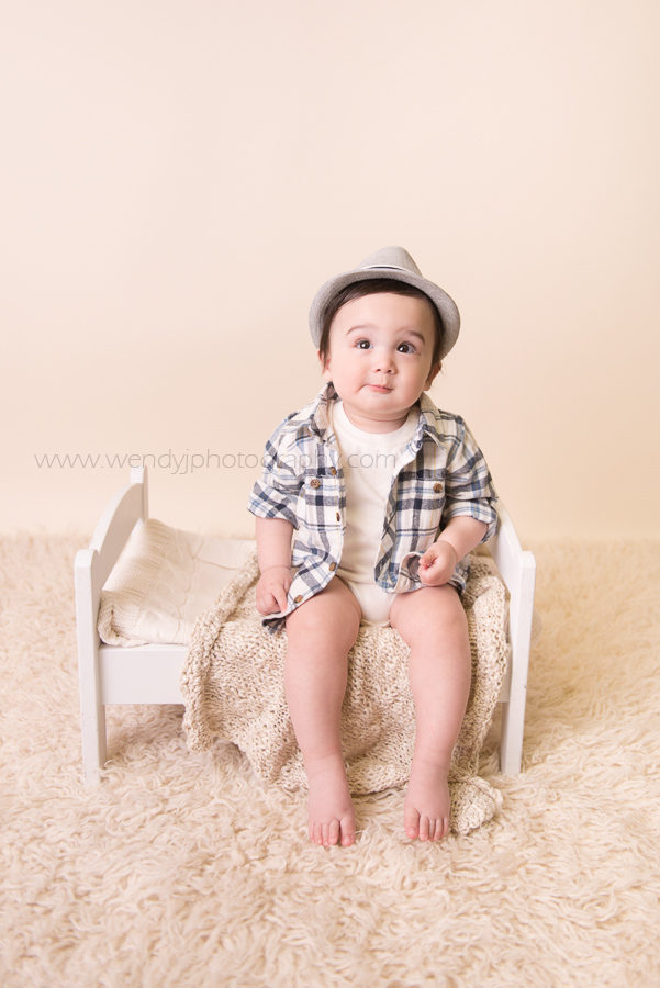 Cute baby boy sits on miniature bed during photo session.