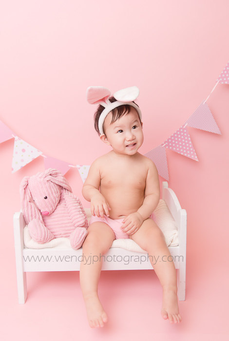 Cute 1 year old girl wearing bunny ears sitting on a miniature bed during a Burnaby, Vancouver B.C. child portrait photography session.